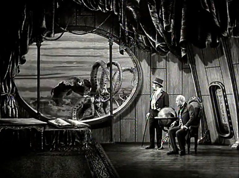 Schwarzweißaufnahme zweier im Stil des 19. Jahrhunderts gekleideter Männer, einer stehend, der andere sitzend, die sich scheinbar in einem U-Boot befinden, vor dessen großem ovalen Fenster eine Oktopus-Figur schwebt