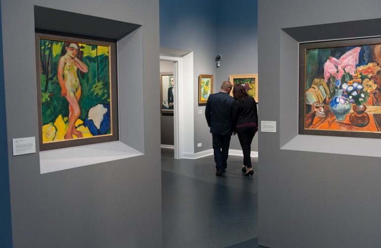 In einem Ausstellungssaal mit großformatigen Gemälden in kräftigen Farben stehen zwei Personen, die in die Kunstbetrachtung vertieft sind.