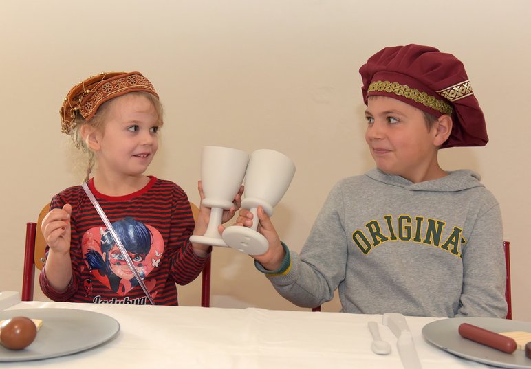 Ein Mädchen und ein Junge mit historischen Kopfbedeckungen sitzen an einem gedeckten Tisch und stoßen mit zwei Kelchen an.