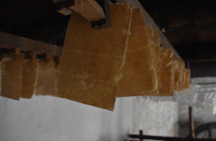 Das Foto zeigt eine große Zahl frisch geschöpfter Blatt Büttenpapier, die zum Trocknen von einer Holzbalkendecke hängen.