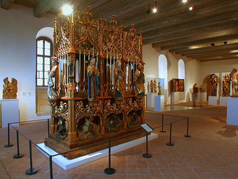 In einem Ausstellungsraum stehen mehrere mittelalterliche Werke sakraler Kunst, im Mittelpunkt ein großer Prunkschrein aus bemaltem und vergoldetem Holz mit einer Vielzahl kunstvoll und detailliert ausgeführter Figuren.
