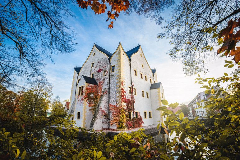 Blick über einen von Herbstlaub umgebenen Wassergraben auf ein kleines, weiß getünchtes Renaissance-Schloss
