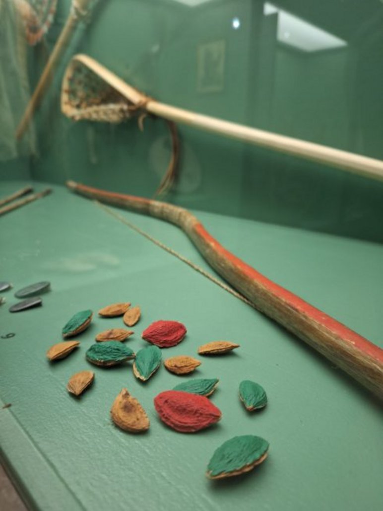 Blick in eine Vitrine, in der rot, grün und blau bemalte Obstkerne sowie bemalte Stöcke und ein Kescher liegen