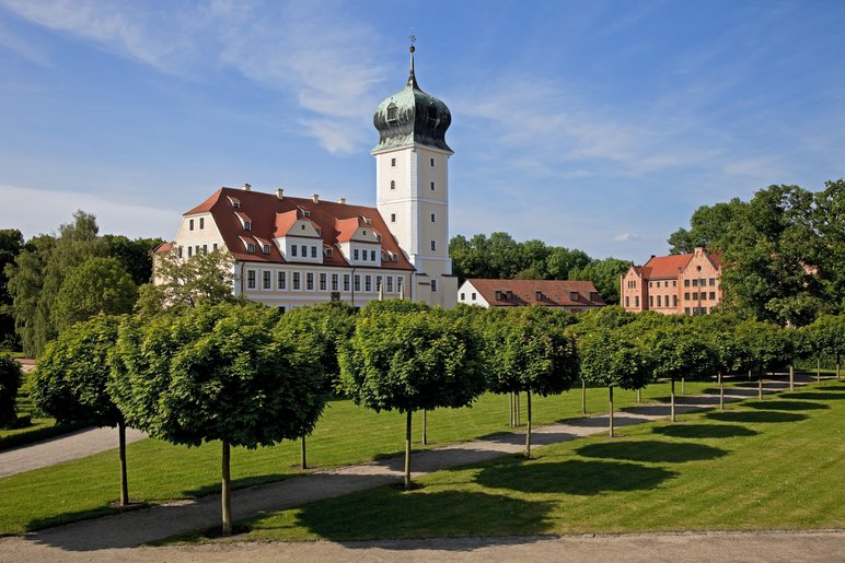 Sommerliche Ansicht eines weißen Schlosses mit prominentem Zwiebelturm in einer gepflegten Parkanlage