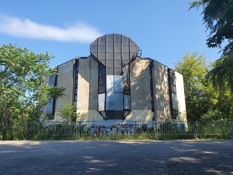 Hinter einer Metallabsperrung ragt, umwuchert von ungemähtem Gras und Bäumen, ein markant gestaltetes Beton-Gebäude auf.