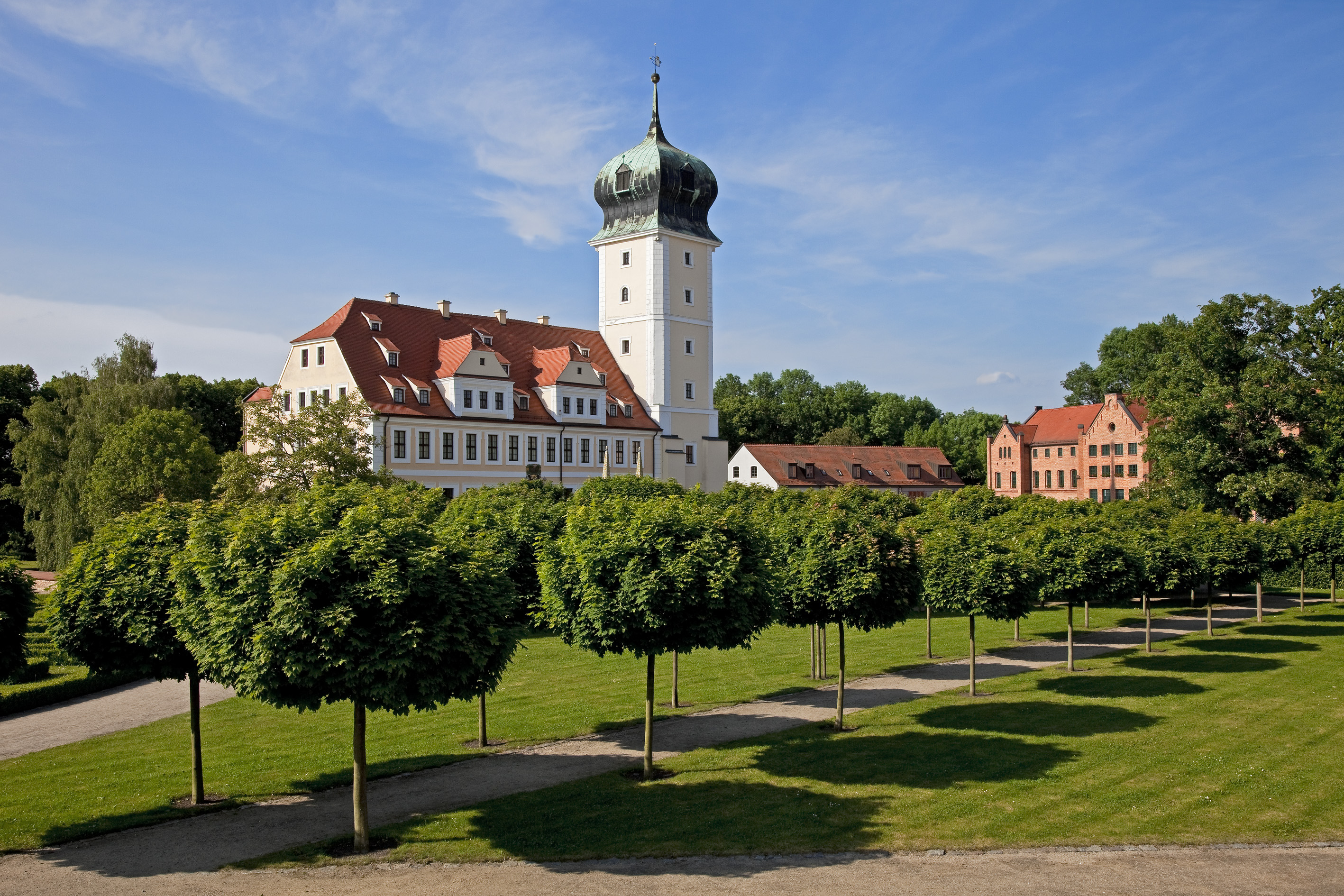 Sommerliche Ansicht eines weißen Schlosses mit prominentem Zwiebelturm in gepflegter Parkanlage