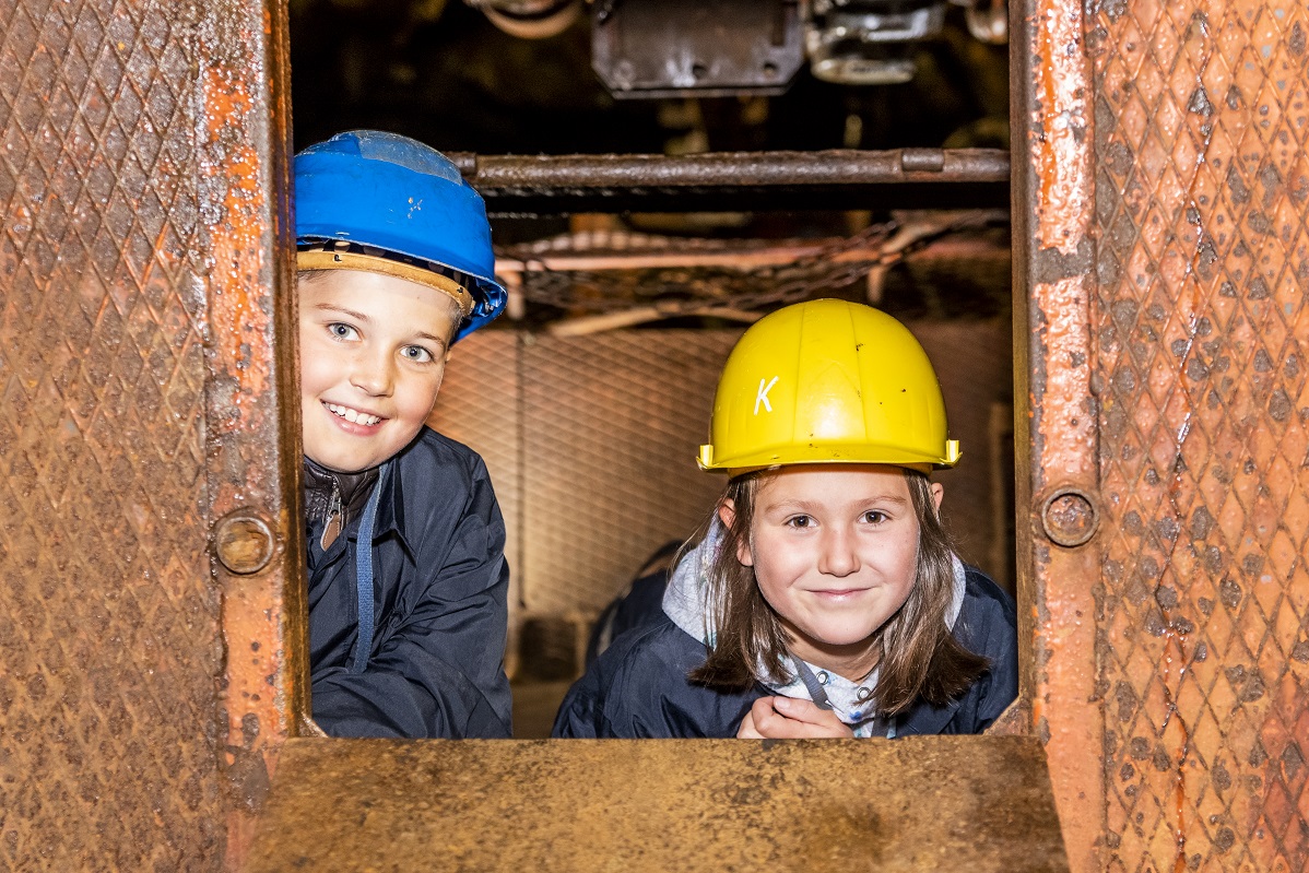 Foto: Zwei Kinder mit Schutzhelm und Schutzumhang schauen aus dem Fenster einer Grubenbahn lächelnd zum Betrachter.